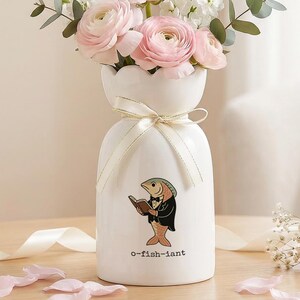 Pode incluir: Um vaso de cerâmica branco com uma ilustração de peixe, segurando flores rosa e brancas. O vaso tem uma fita amarrada ao redor do pescoço e o texto "o-fish-iant" na frente. O vaso está em uma mesa de madeira.