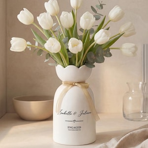 Könnte beinhalten: Eine weiße Keramikvase mit gewelltem Rand, die einen Strauß weißer Tulpen und Eukalyptus enthält. Die Vase hat ein goldenes Band und den Text "Isabelle & Julian" und "ENGAGED". Eine beige Schüssel und eine kleine Glasvase sind ebenfalls sichtbar.