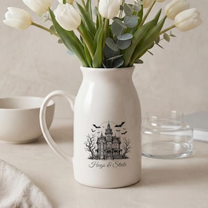 Może przedstawiać: Biały ceramiczny dzbanek z czarną grafiką przedstawiającą nawiedzony dom, nagie drzewa i nietoperze z napisem Hugo & Strile. Dzbanek wypełniony jest bukietem białych tulipanów i zielonymi gałązkami eukaliptusa.