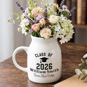 Op de afbeelding: Een witte keramische vaas met een handvat, gevuld met een kleurrijk boeket bloemen. De vaas is beschreven met "CLASS OF 2026" en een naam, perfect voor een afstudeercadeau. De vaas staat op een houten tafel.