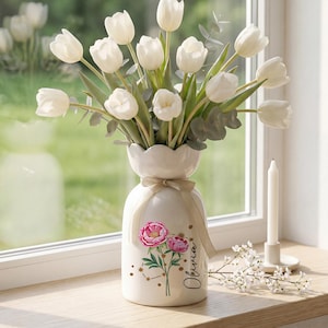 Puede incluir: Un jarrón de cerámica blanco con un diseño floral y el nombre "Olivia" en escritura cursiva, con tulipanes blancos y eucalipto. Una vela blanca y pequeñas flores blancas están en un alféizar de madera. El jarrón está atado con una cinta beige.