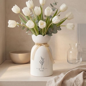 Puede incluir: Un jarrón de cerámica blanco con borde festoneado contiene un ramo de tulipanes blancos y eucalipto. El jarrón tiene un diseño botánico negro y una cinta dorada atada alrededor del cuello. El texto "J&K 02.15.20" está impreso en el jarrón.