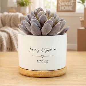 Peut inclure: Un pot de fleurs en céramique blanche avec une plante succulente, posé sur une base en bois. Le pot porte les noms "Henry & Sophia", un cœur et la date "06.20.2026". La succulente a des feuilles gris-vert.