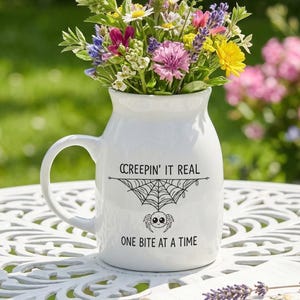May include: A white ceramic pitcher-style mug filled with a colorful bouquet of wildflowers. The front of the mug features a black graphic of a spider hanging from a web with the text "CREEPIN' IT REAL" above and "ONE BITE AT A TIME" below.