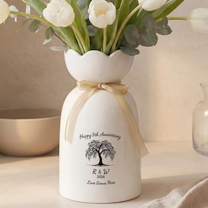 Puede incluir: Un jarrón de cerámica blanca con borde festoneado contiene un ramo de tulipanes blancos y eucalipto. El jarrón tiene una cinta atada alrededor del cuello y presenta el texto "Happy 5th Anniversary" y "Love Grows Here."