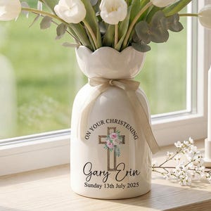May include: A white ceramic vase with a floral arrangement of white tulips and greenery. The vase has a cross and floral design with the text "ON YOUR CHRISTENING" and personalized names and date. A white candle and flowers are on the windowsill.