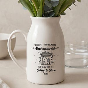 May include: A white ceramic pitcher with a handle, filled with white tulips and greenery. The pitcher has a black graphic of a house and text that reads "New memories" and "178 Chestnut IT Calley & Steve 2025".