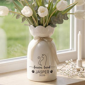 Puede incluir: Un jarrón de cerámica blanca con borde festoneado, atado con una cinta beige, contiene tulipanes blancos y vegetación. El jarrón está inscrito con una silueta de gato, las palabras "forever loved" y el nombre "Jasper" con el año 2026.