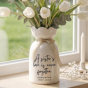 Pode incluir: Um vaso de cerâmica branca com borda ondulada contém tulipas brancas e vegetação. O vaso tem uma fita bege e o texto "O amor de uma irmã nunca é esquecido" com um nome e datas. Uma vela branca está por perto.