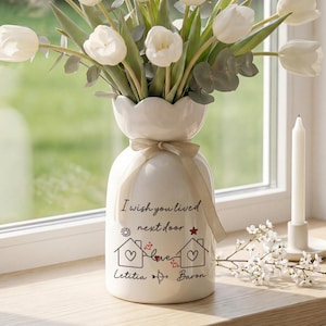 Op de afbeelding: Een witte keramische vaas met een geschulpte rand bevat witte tulpen en groen. De vaas heeft een crèmekleurig lint en de tekst "I wish you lived next door". Een witte kaars staat in de buurt op een houten vensterbank.
