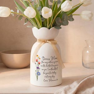 May include: A white ceramic vase with a scalloped top, holding white tulips and eucalyptus. The vase has a floral design and text that reads "Bonus Mom". A beige ribbon is tied around the vase.