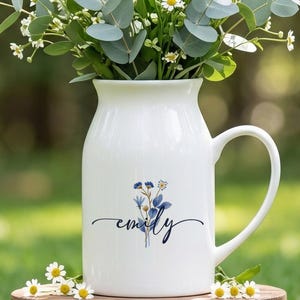 Pode incluir: Um jarro de cerâmica branca com pega, apresentando um gráfico floral azul e o nome "emily" em letra cursiva. O jarro está cheio com um ramo de margaridas brancas e folhas de eucalipto, pousado sobre um pequeno banco de madeira.