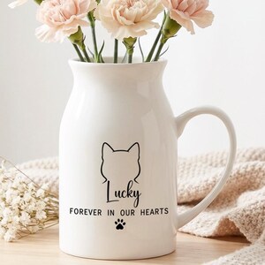 May include: A white ceramic pitcher vase featuring a black line drawing of a cat head, the name Lucky, the text FOREVER IN OUR HEARTS, and a small paw print. The vase is filled with light pink carnations and sits next to small white flowers.