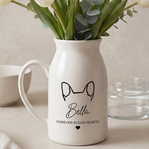May include: A white ceramic pitcher vase with a black dog silhouette, the name "Bella", and the text "Forever in our hearts". The vase holds a bouquet of white tulips and greenery. A small white bowl and a glass vase are also visible.