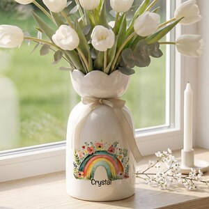 Puede incluir: Un jarrón de cerámica blanca con borde festoneado y una cinta beige atada alrededor del cuello. El jarrón presenta una colorida ilustración de arcoíris rodeada de pequeñas flores y el nombre Crystal. Contiene un ramo de tulipanes blancos y eucalipto.