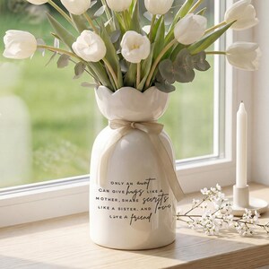 May include: A white ceramic vase with a scalloped rim holds white tulips and greenery. The vase has a beige ribbon and text that reads "ONLY AN AUNT CAN GIVE HUGS LIKE A MOTHER, SHARE SECRETS LIKE A SISTER, AND LOVE LIKE A FRIEND."