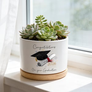 Pode incluir: Um vaso de cerâmica branco com tema de formatura, com um capelo, um diploma e o texto "Congratulations On your Graduation". O vaso contém várias suculentas verdes e está sobre um porta-copos de madeira.