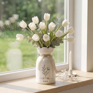 Puede incluir: Un jarrón de cerámica blanca con borde festoneado contiene tulipanes blancos y vegetación. El jarrón tiene un diseño floral con las iniciales "R & M" y la fecha "07.10.26". Una vela y pequeñas flores están cerca.