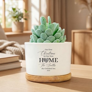 Pode incluir: Um vaso de cerâmica branco com uma planta suculenta verde. O vaso tem o texto "Our First Christmas In Our New HOME The Smiths 2831 Greenleaf Ave. 2021" em preto. Ele está sobre um descanso de madeira redondo marrom claro.