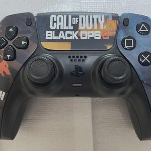 May include: A black and gray PlayStation 5 controller with a "Call of Duty: Black Ops 6" design. The controller features black buttons, analog sticks, and a PlayStation logo. The design includes orange and blue accents with the words "Truth Lies".