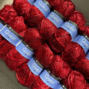 May include: A collection of red yarn skeins, labeled "Quest" by Berroco. The yarn is a vibrant, deep red color, and the labels are blue with white text. The skeins are arranged closely together, filling the frame.