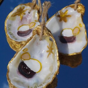 Può includere: Tre ornamenti a conchiglia dipinti a mano con bordi dorati e ganci in spago. Ogni conchiglia presenta una scena della natività con una stella e il testo "Isaia 9:6". Le conchiglie sono esposte su una superficie riflettente.