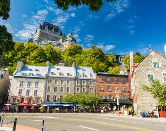 May include: A view of a historic neighborhood in Quebec City, Canada. The street is lined with stone buildings, some with red tile roofs. A funicular railway is visible in the background, leading up to a large castle-like building on a hill.