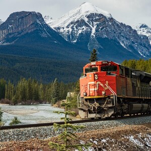 May include: A red freight train with a white and black logo travels through a mountain valley. The train is traveling on a track with a river and snow-capped mountains in the background.