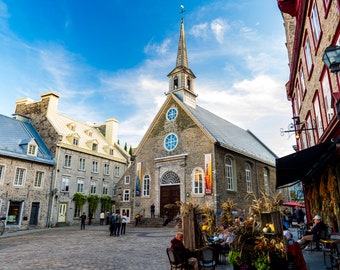 May include: A gray stone church with a tall steeple and a round window. The church is located in a city square with other buildings and people walking around.