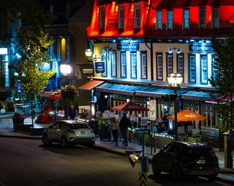 Puede incluir: Una escena nocturna de una calle con un restaurante en el lado derecho de la imagen. El restaurante tiene un toldo rojo y un letrero que dice "A Bistro du Vieux Quebec". Hay mesas y sillas dispuestas fuera del restaurante, y la gente está sentada en las mesas. Hay dos coches aparcados en la calle, uno blanco y otro negro.