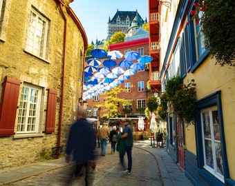 Puede incluir: Una calle estrecha de adoquines en una ciudad con edificios coloridos a ambos lados. Paraguas a rayas azules y blancas cuelgan por encima, creando un dosel de color. La gente camina por la calle, disfrutando del día soleado.