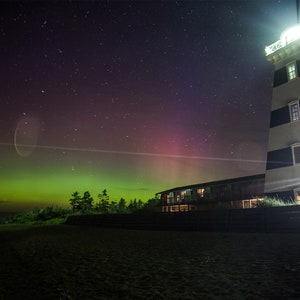 May include: A black and white striped lighthouse with a bright light shining from the top. The lighthouse is on a beach with a dark sky filled with stars and a green and pink aurora borealis in the background.