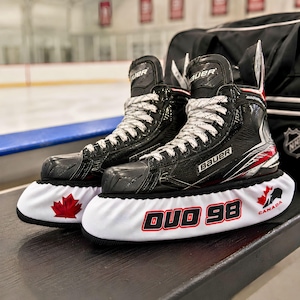 Puede incluir: Un par de patines de hielo negros Bauer con cordones blancos, cada uno con un protector de cuchilla blanco. Los protectores presentan una hoja de arce roja, el texto DUO 98 en rojo y negro, y un pequeño logotipo de Canadá con una hoja de arce.