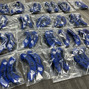 以下が含まれることがあります： 透明なビニール袋に個別に包装された、複数の青いスポーツサポート。サポートには白いアクセントとテキストがあります。商品は表面に配置されています。