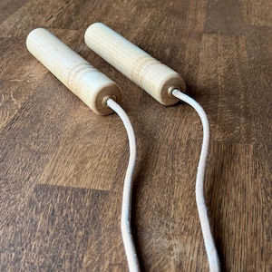 Può includere: Una corda per saltare in legno con manici in legno color chiaro e una corda color crema. I manici cilindrici hanno un raccordo metallico color ottone dove è attaccata la corda. Lo sfondo è una superficie di legno scuro.