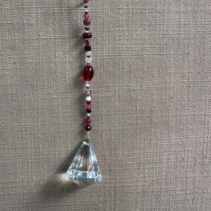 Puede incluir: Un atrapasoles decorativo de cristal en forma de prisma con un lazo de metal dorado. El atrapasoles presenta una cadena de cuentas rojas, rosas y transparentes. El prisma es de cristal transparente y facetado.