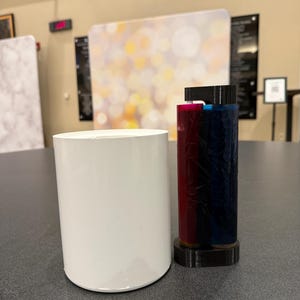 May include: A white cylindrical object and a cylindrical object with red, blue, and black elements sit on a gray surface. The objects are in front of a blurred background with a light and dark color scheme.