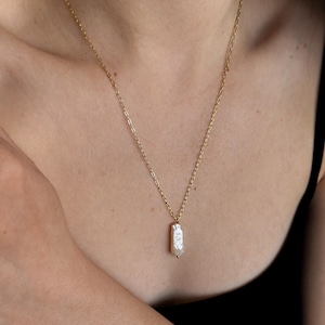 May include: A gold chain necklace featuring a rectangular, off-white pearl pendant. The pendant has a textured surface and a small gold bead at the base. The necklace is set against a dark backdrop.