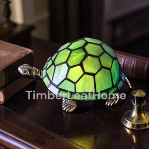 Peut inclure: Lampe décorative en forme de tortue avec une carapace en vitrail vert et noir. La lampe a des pieds, une tête et une queue de couleur bronze. Le texte "TimberLeafHome" est imprimé sur la surface.