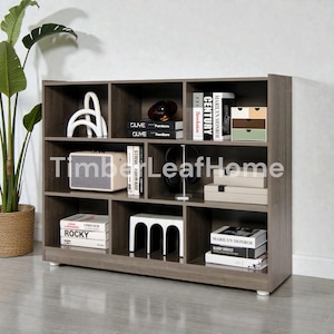 Peut inclure: Une bibliothèque en bois gris avec plusieurs compartiments, remplie de livres, d'objets décoratifs et d'un petit haut-parleur. La bibliothèque est de forme rectangulaire et est placée contre un mur blanc. Le texte "Timber Leaf Home" est visible.
