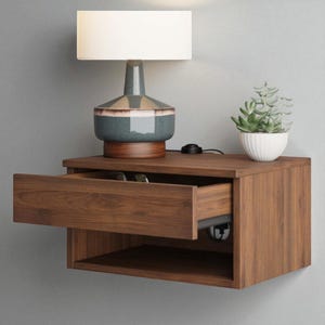 May include: A floating wooden nightstand with an open drawer, a lamp, and a small potted plant. The nightstand is made of dark wood and has a rectangular shape. The lamp has a white lampshade and a decorative base. The plant is a small succulent in a white pot.