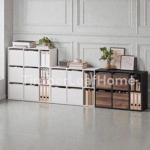 Peut inclure: Ensemble de meubles de rangement modulaires blancs et noirs. Les unités blanches ont des compartiments carrés avec des portes fermées, tandis que l'unité noire comporte des étagères ouvertes et des tiroirs en bois.