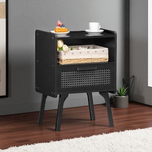 May include: A black nightstand with a woven drawer and a shelf. A white teacup and a plate with a pastry sit on top. A basket and flowers are on the middle shelf. The nightstand has four angled legs.