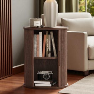 May include: A dark brown, semi-circular side table with a built-in bookshelf. The table features a top shelf with a white vase and a small candle. The bookshelf holds several books and a vintage camera.