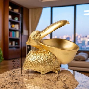 May include: A gold-colored pelican sculpture with an open beak, designed to hold items. The sculpture is 9 inches (22.8 cm) tall and 8.7 inches (20.3 cm) wide. The pelican's feathers are detailed, and the sculpture is placed on a marble-like surface.