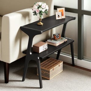 May include: A black two-tiered side table with a unique, modern design. The top shelf holds a vase of flowers and framed photos. The lower shelf displays a vintage camera, books, and a small wooden box. A woven basket sits below.