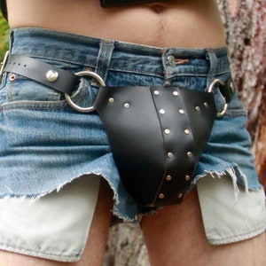 Puede incluir: Una prenda de cuero negro con remaches plateados y un cinturón, usada sobre unos shorts vaqueros. Los shorts son azules con forros de bolsillo blancos y un dobladillo deshilachado. La prenda tiene dos grandes anillos plateados sujetos al cinturón.