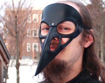 May include: A person wearing a black masquerade mask with a long, pointed beak. The mask has cutouts for the eyes and nose. The person is wearing a black leather jacket.