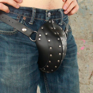 May include: A black leather codpiece featuring a rounded, molded shape adorned with numerous silver-toned metal studs. The codpiece is attached to a black leather belt with silver-toned hardware, worn over a pair of blue denim jeans.