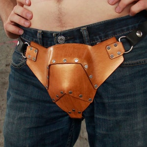 May include: A brown leather belt with metal studs and a buckle. The belt is worn around the waist and has a large, decorative piece attached to the front.